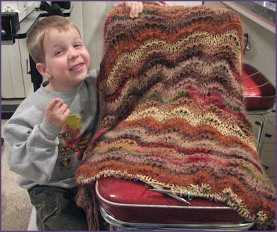 boy smiles with goofy grin, posing next to afghan in progress